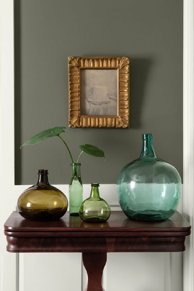 A hallway vignette featuring a console table decorated with colourful glass vases.  The upper walls are painted in Dark Olive 2140-30 and the woodwork and panelling features Icicle OC-60.