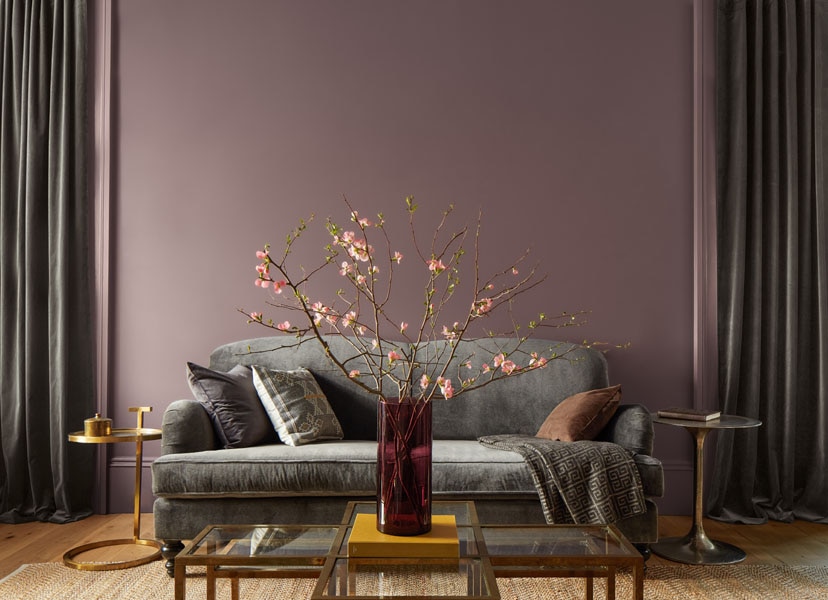 A living room colour drenched in quietly colourful Cinnamon Slate 2113-40 to infuse a sense of elevated ease. The room is styled with a grey sofa and large floral arrangement.