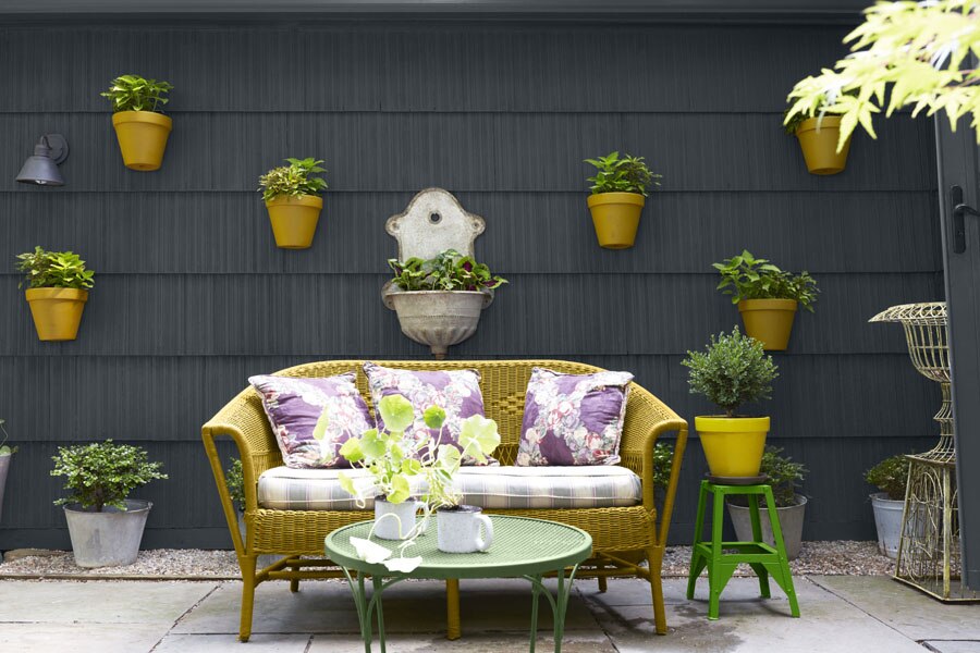 A garden patio with an ochre coloured rattan sofa and planters, featuring a wooden clad wall painted with Aura Exterior Low Lustre in Raccoon Fur 2126-20