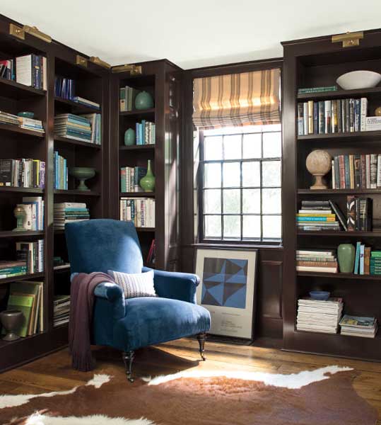 A library feauturing floor to ceiling bookcases painted in Tarpley Brown CW-170. The room is styled with a blue velvet accent chair and striped blind window dressing